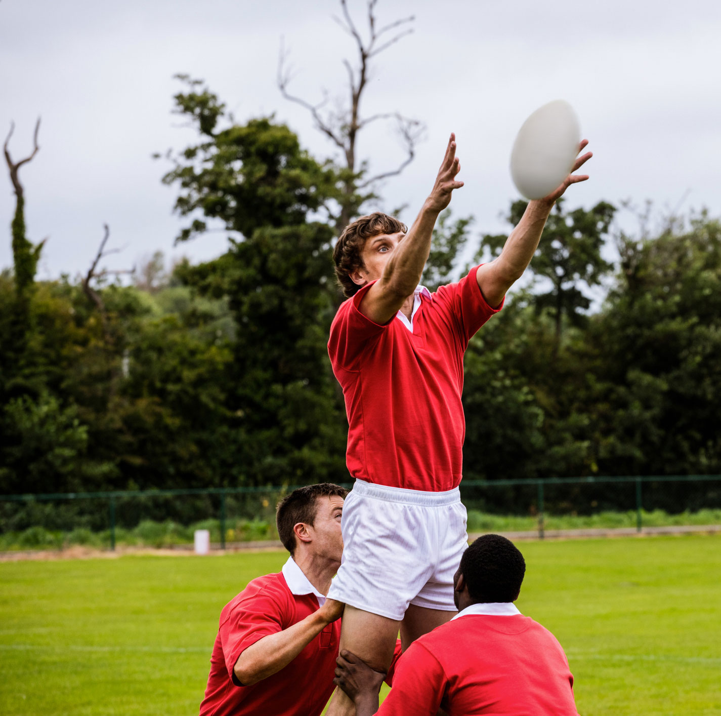 Male rugby players in a red kit with white shorts take a lineout 