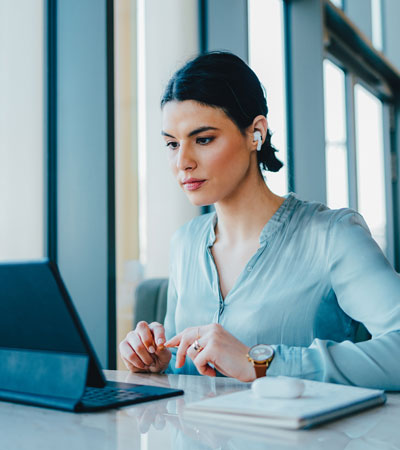 Beautiful businesswoman sitting at the desk, looking down at her tablet while wearing earbuds. She is having a video call with someone.