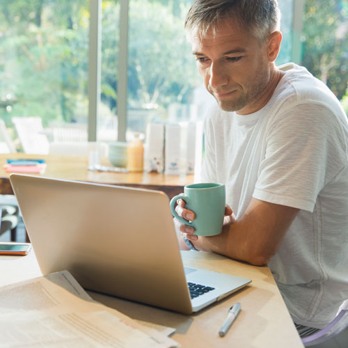 Man in casual wear sitting at the breakfast table on his laptop while drinking coffee
