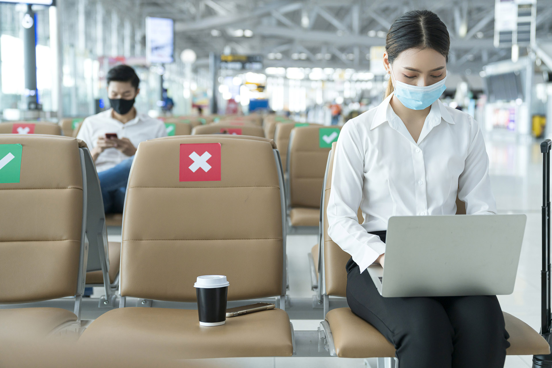 Social distancing, businesswoman sit working with laptop and smartphone keeping distance away from each other to avoid covid19 infection during pandemic. Empty chair seat red cross shows new normal travellers using their smart devices in the airport