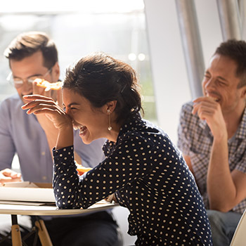 Indian woman laughing eating pizza with diverse coworkers in office Marketing Consultant