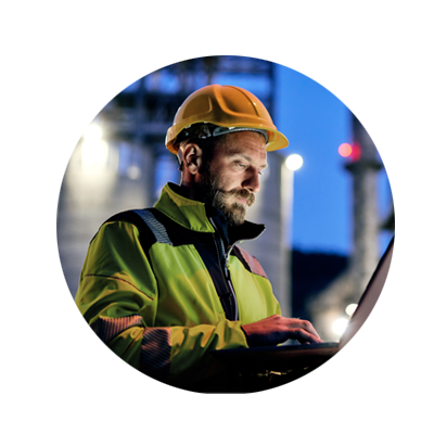 Man in a high-vis jacket and orange hardhart standing outside a power plant of an evening while holding a laptop 