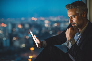 Businessman sitting on a windowsill at night and looking at a digital tablet, with cityscape at the background and copy space.